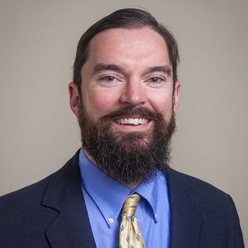 Image of Burkett CPAs Vice President Matthew Hodges, smiling, bearded, wearing a blue suit, blue shirt and yellow tie