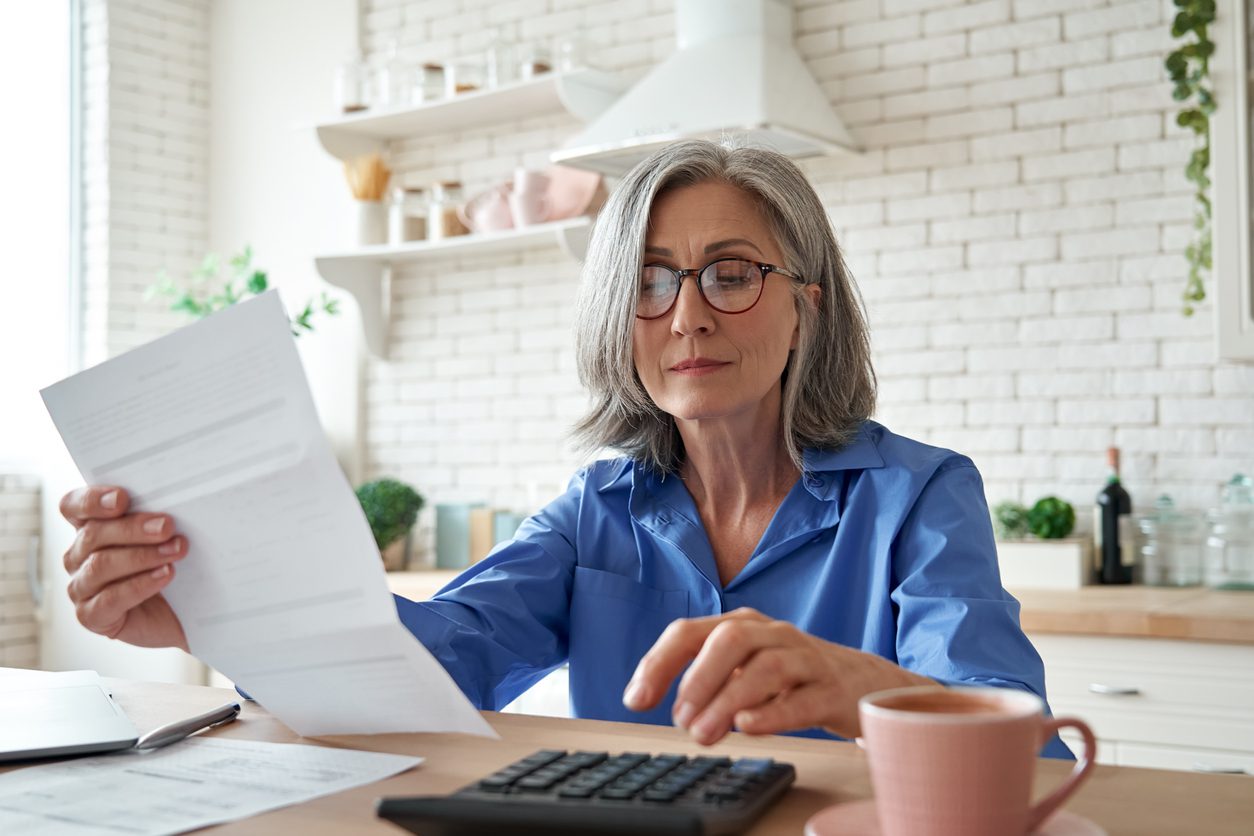 older woman reads tax or retirement document while making calculations on a desk calendar