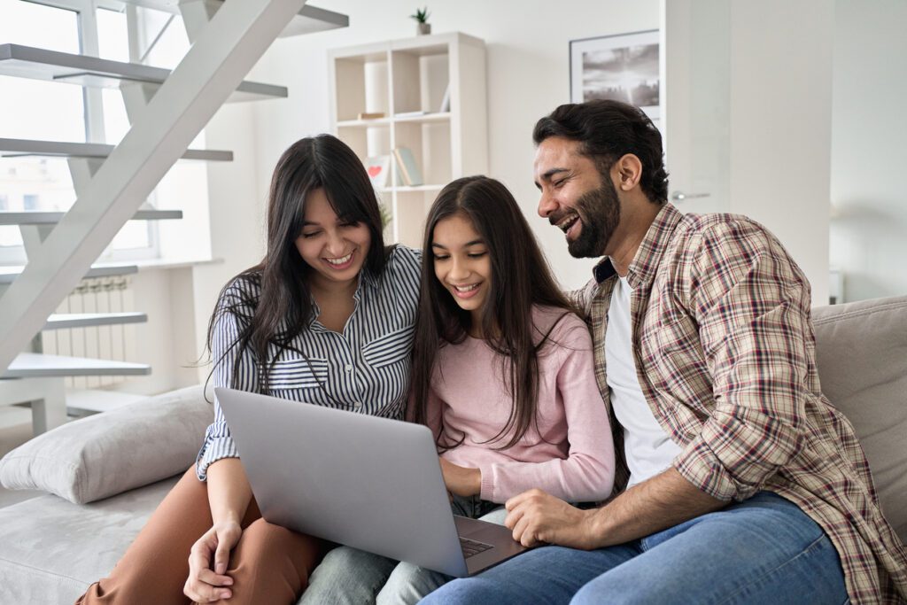 Happy family with child daughter having fun using laptop computer at home. Smiling parents and teen kid daughter laughing looking at device browsing or watching funny videos sitting on sofa.