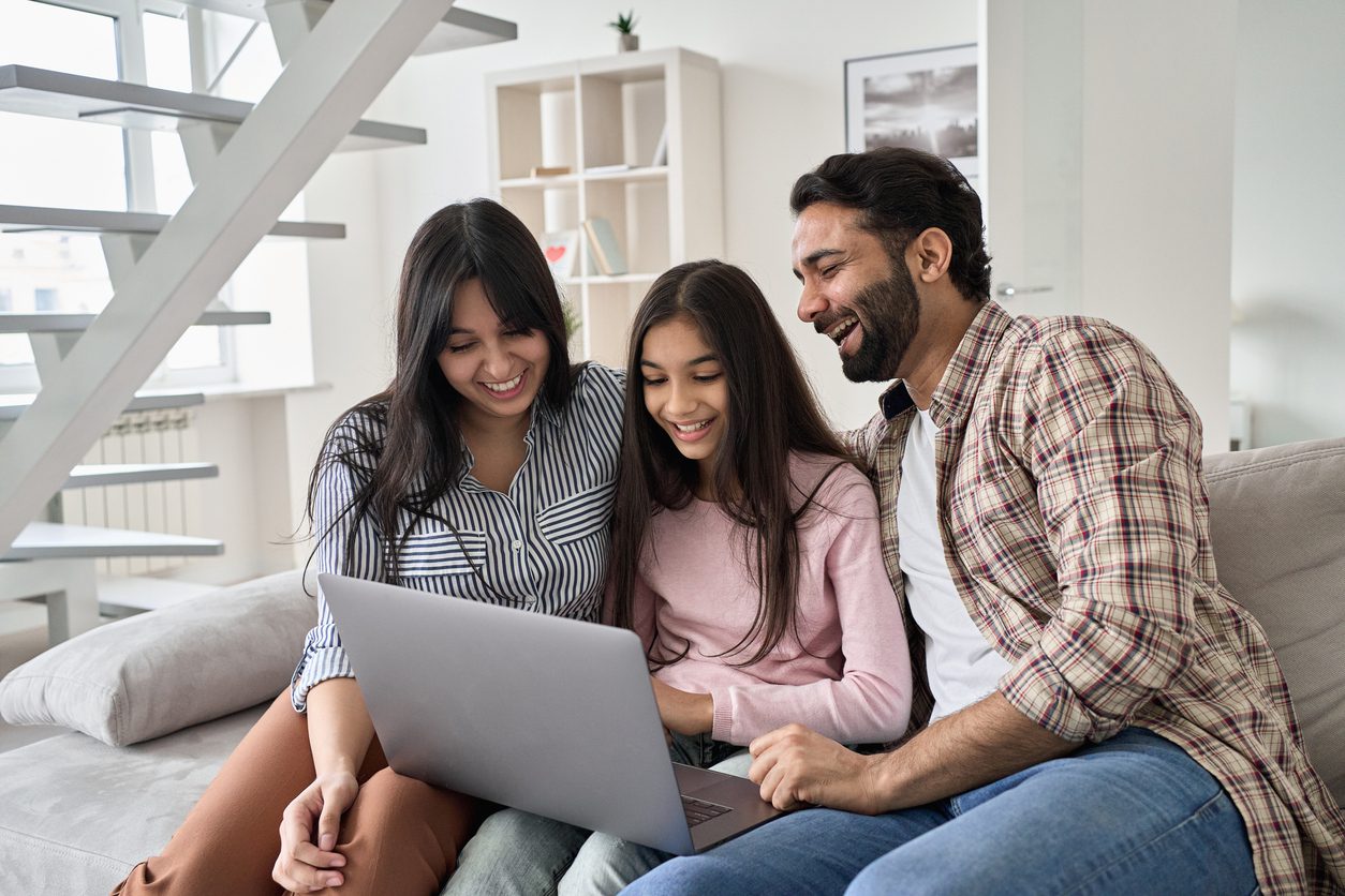 Happy family with child daughter having fun using laptop computer at home. Smiling parents and teen kid daughter laughing looking at device browsing or watching funny videos sitting on sofa.